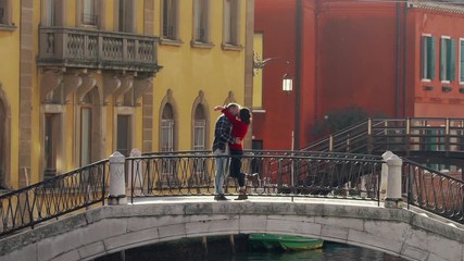 Handsome loving man kissing and hugging his charming asian girlfriend standing on the ancient Venetian bridge during their honeymoon. Travelling to Europe as the best way of spending the vacation.