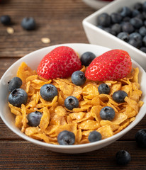 Corn flakes with blueberries and raspberries in white bowl in dark wooden desk.