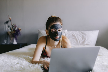 Young lady with clay mask browsing social media on laptop while resting on comfortable bed in cozy bedroom