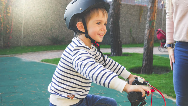 Toned Portrait Of Cheerful Smiling Little Boy In Black Protective Helmet Riding His Bicycle