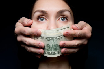 The mouth of a young woman is closed by a stack of dollar bills. Buying silence