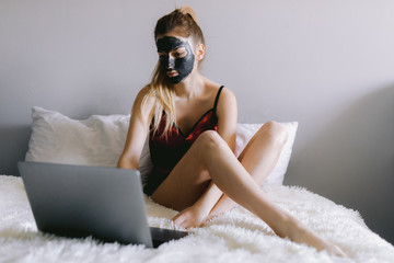 Young woman in pajama and with clay mask on face sitting on bed and watching video on laptop during spa routine at home