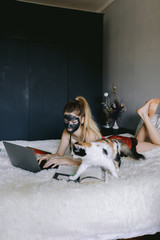 Young woman in nightwear and mud mask lying on bed near cat and doing homework assignment on laptop