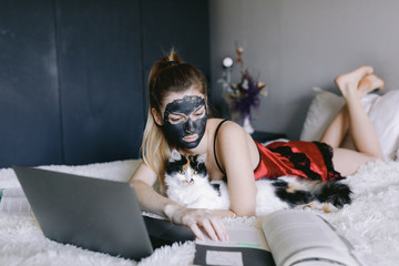 Young woman in sleepwear and clay mask hugging furry cat and reading textbook while lying on bed near laptop and doing homework