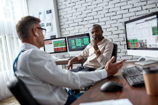 Ensuring Finacial Freedom. Two diverse colleagues traders talking to each other while sitting in the office in front of multiple computer screens.