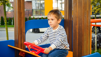 Happy smiling boy playing on children playground at park and turning steering wheel of wooden car