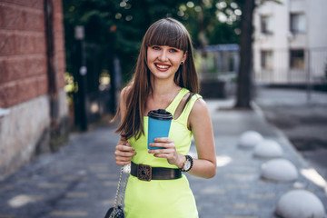 Pretty brunette girl in green dress with cup of coffee walking outdoor on the street