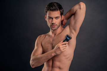 Handsome young bearded man isolated. Portrait of topless muscular man is standing on gray background with electric Shaver in hand while shaving armpits. Men care concept.