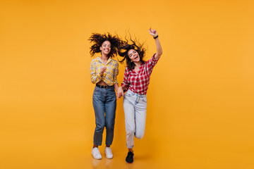 Positive girls jump on orange background. Full length shot of best friends in denim pants