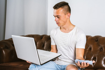 A young man sitting on a sofa in one hand holds a computer in the other hand a face mask, weighs what to choose, works remotely during quarantine, pandemic COVID-19, self-isolation. Virus