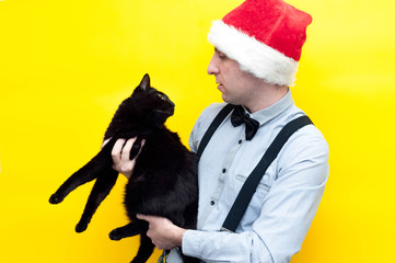 handsome man in red christmas santa hat holding and looking at cute scared big fat black cat on yellow background with copy space