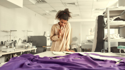 Excellence and Quality. Young female clothing designer leaning against the desk with fabric,...