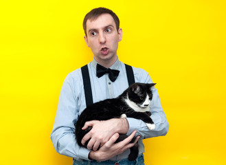 handsome man with face expressions in blue shirt holding adorable black and white taxedo cat and looking away on yellow background with copy space