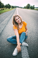 Fototapeta premium A young girl in a yellow T-shirt and blue jeans and white sneakers sits on the road and smiles. Evening, soft light. Light noise in the photo. Setting goals and moving towards them