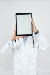 Male doctor in medical uniform with stethoscope around neck holding blank digital tablet in front of his face, standing against grey background