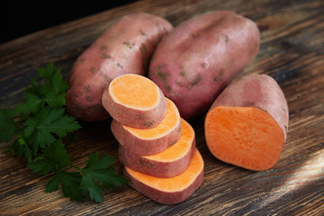 Raw sweet potatoes (batatas) on a rustic wooden table decorated with parsley. One batata in the front is half sliced. Slices are stacked like a ladder (stairs). Top view