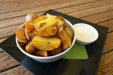 Fried potato wedges in a bowl and a garlic dip on a black tray on a rustic wooden table, copy space