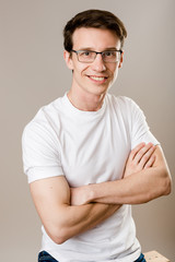 Young, confident production worker wearing glasses and a shirt sits on a grey background with his hands crossed on his chest and a nice, trusting smile looking at the camera.