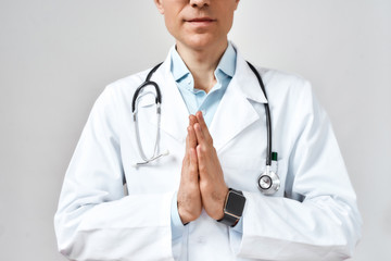 Cropped photo of male doctor or physician in white coat with stethoscope around his neck praying to God for help while standing against grey background