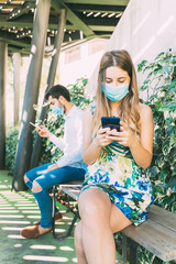 young couple maintaining social distance in a bank, both wearing a medical mask and using their phone.