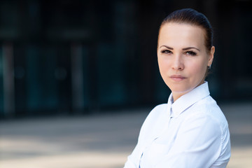 Beautiful confident young woman, successful girl businesswoman in white formal shirt standing outdoors near business centre, look at camera. Copy space, place for text. Portrait of serious strict lady