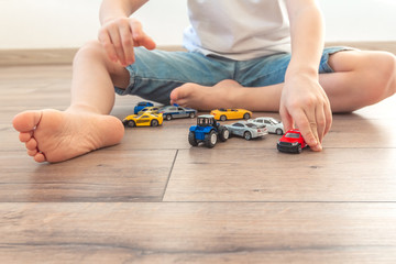 Little boy is playing with colored toy cars at home. Children feet barefoot on wooden warm floor. Joyful toddler sitting in room on laminate with  many favorite toys. Happy childhood