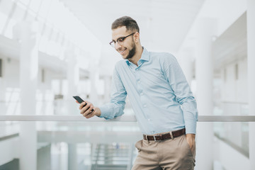 University student wearing blue shirt and glasses looking at his smartphone standing in a bright corridor