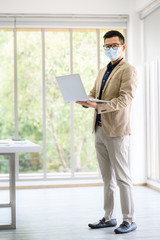 Fototapeta premium Happy Asian businessman entrepreneur wears face mask standing and holding laptop with confident in meeting room at office