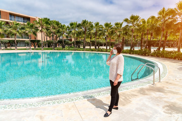 A middle-aged woman, on a sunny day, in a protective mask against the virus, coronavirus, allergies, talking on the phone near the pool in the hotel.