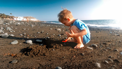 Little toddler boy playing on the sea beach with plastic shovel and building sand castle