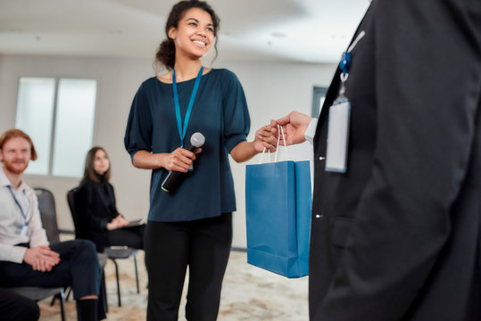 Breaking Down Barriers. Young Female Speaker Receiving Gift Bag From Her Male Colleague At Business Meeting, Forum. Audience Sitting At The Conference Hall