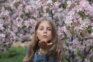 Fototapeta premium Portrait of a young girl in a flowering garden who sends an air kiss. The girl has long blond hair. The trees are beautiful pink flowers.