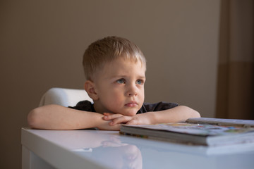 Naklejka premium Portrait of a little boy with blond hair who sits at a table and puts his head on his hands