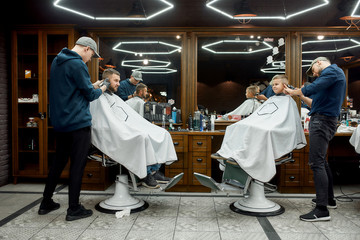 Family at barbershop. Young father and his little happy son sitting together in chairs in the...