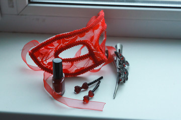 Composition of red vintage carnival mask, nail polish, metal hair clips with rhinestones, pomegranate beads made of natural stone on a white background on the windowsill.