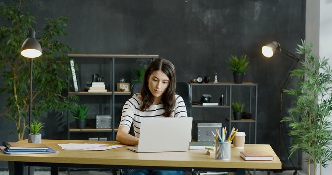 Young Caucasian busy female office worker sitting at table, working on laptop computer and considering.
