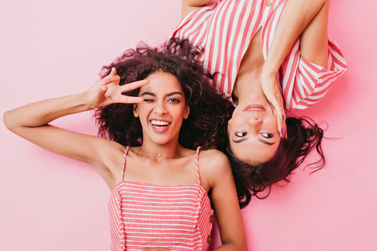 Professional photo in studio of young and attractive girls with beautiful makeup and dark curly hair. Women are lying on floor, fooling around and showing peace sign