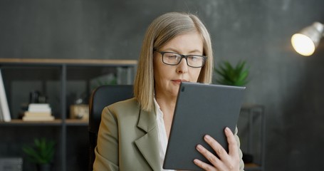 Beautiful senior woman in glasses tapping and scrolling on computer in office.