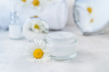 Obraz na płótnie Canvas Herbal cosmetic cream in opened glass container and fresh chamomile flowers, towel, mirror on a white background. Natural organic moisturizer skincare product. Selective focus. Copy space.