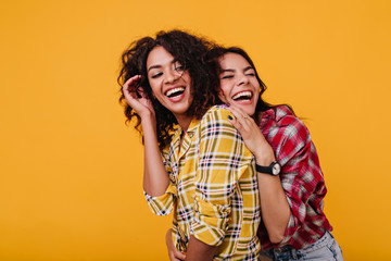Girls in stylish street clothes sincerely laugh at each other's jokes. Girlfriends happily pose for common photo in studio - obrazy, fototapety, plakaty