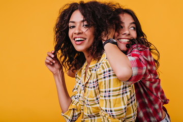 Female students with tanned skin are fooling around on orange isolated background. Pretty girl tousling her friend's hair