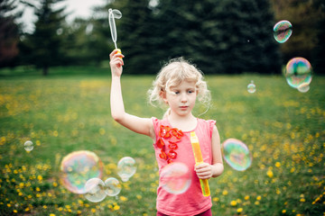 Preschool Caucasian blonde girl blowing soap bubbles in park on summer day. Child having fun outdoors. Authentic happy childhood magic moment. Lifestyle seasonal activity for children.