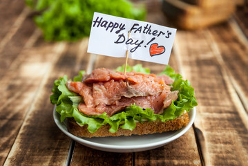 Red fish sandwich on rye bread with lettuce leaf on a dark wooden background. A note of congratulations.