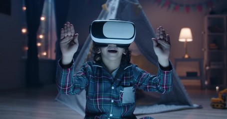 Funny little kid wearing pajamas is putting on virtual reality headset to dive into video games before bedtime , getting lit and having fun 4k footage - Powered by Adobe