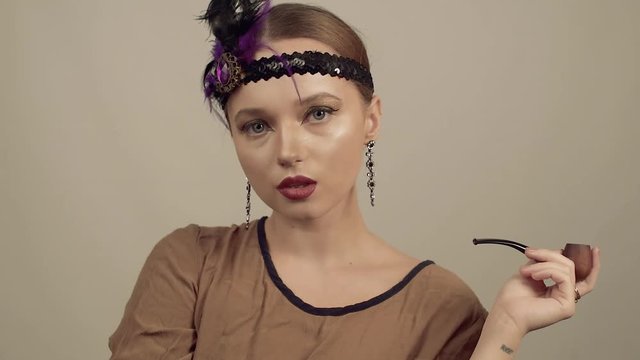 Girl in the 90s style, chicago style. Hat, headband with decorative feathers on the head. Incredible jewelry, long earrings with diamonds, coquette. Lady smokes a pipe