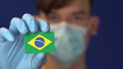 COVID-19 pandemic in Brazil. A young, attractive Brazilian Latin American man in protective medical mask and gloves holding the flag of Brazil. Coronavirus pandemic in South America - Powered by Adobe