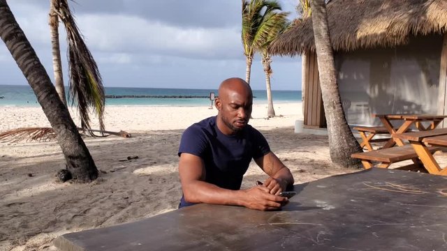 Black African American millennial  man on the beach checks his mobile phone to read the news, 4k  