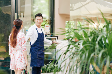 Cheerful young Vietnamese cafe owner in denim apron welcoming female client in his cafe