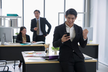 Happy asian young handsome smart successful businessman so excited he looking reading good news at his mobile phone with team mates discussing in the sunset background at workplace