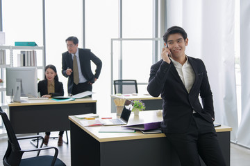 Happy asian young handsome smart successful business man smiling while received good news from mobile phone with team mates discussing in the background at workplace